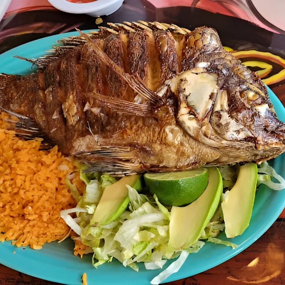 A whole fried fish with rice, shredded lettuce, avocado slices, and lime on a blue plate.