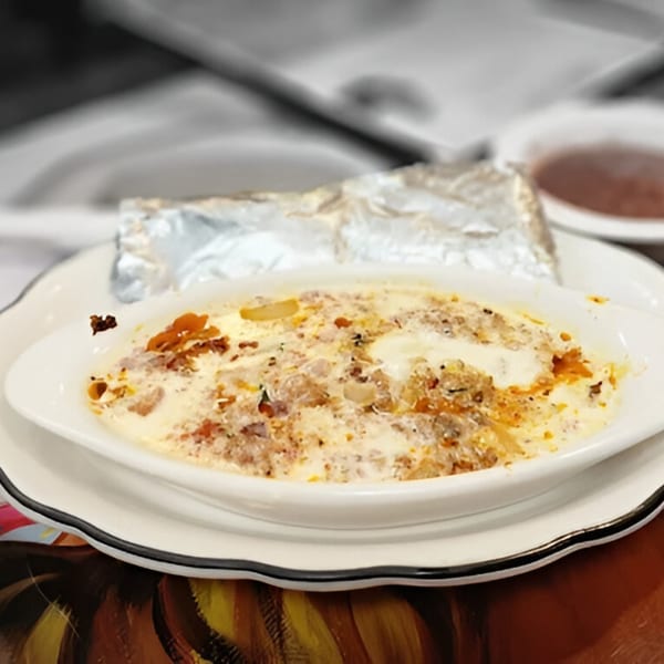 A white oval dish of creamy, cheesy Mexican food with a foil-wrapped item and sauce in the background.