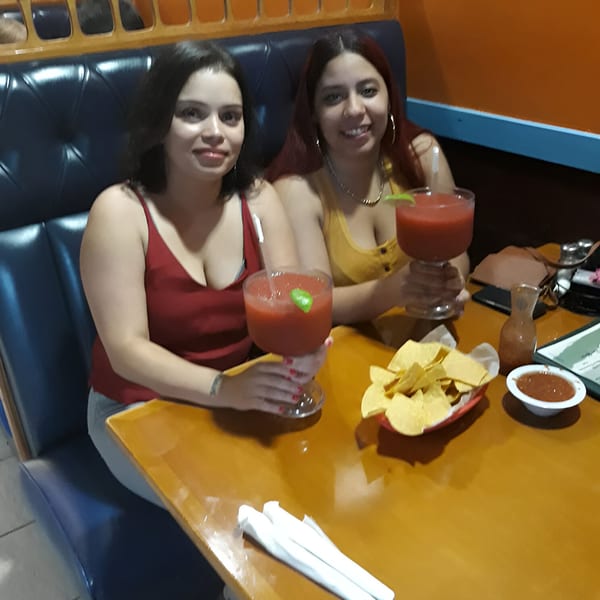 Two women smiling, holding red margaritas, with chips and salsa on a table at Los Gallos Mexican Grill.