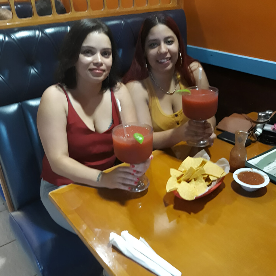 Two women smiling, holding red margaritas, with chips and salsa on a table at Los Gallos Mexican Grill.
