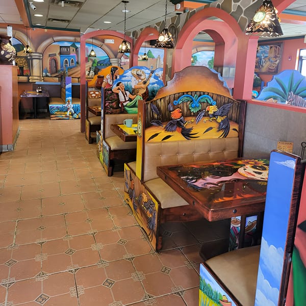 Colorful Mexican restaurant interior with painted booths, tables, and murals depicting village scenes and roosters.
