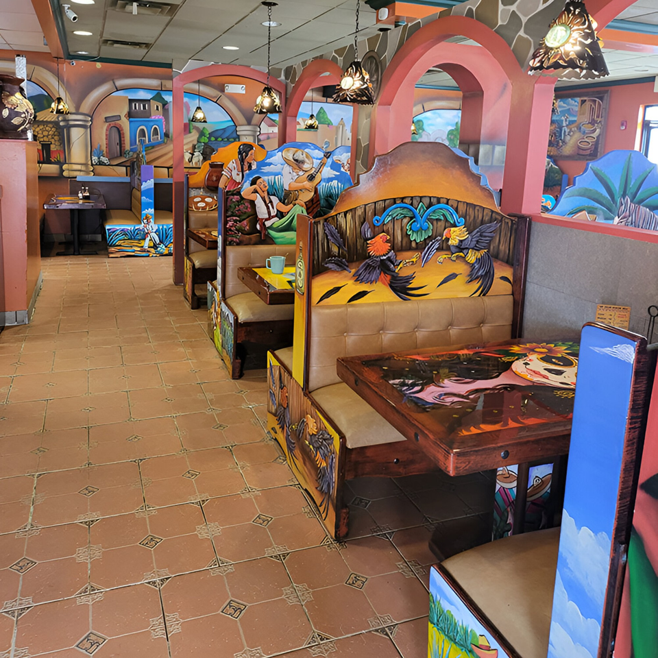 Colorful Mexican restaurant interior with painted booths, tables, and murals depicting village scenes and roosters.